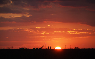 [One-third of the yellow-orange orb sits above the dark horizon. The outline of an oil derrick and three storage tanks as well as some trees are visible. The clouds in the immediate background of are shades of orange.]